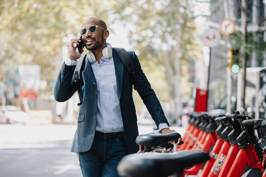 Happy businessman talking on smart phone near bicycles on street