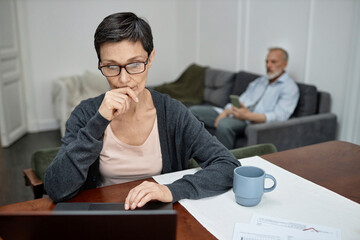 Woman using laptop working from home husband sitting on sofa in background