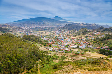 A scenic panorama of Tenerife with verdant hills, a town with mixed architecture, and the majestic Mount Teide rising in the background