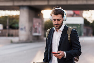Young businessman wearing headphones and using smart phone at street