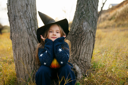 Cute girl in witch costume sitting by tree
