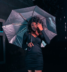 A woman is holding a silver umbrella and wearing a black dress. The umbrella is open and the woman...