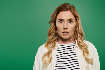 Fototapeta premium Curly-haired woman in casual attire expresses surprise while standing on a bright green background.