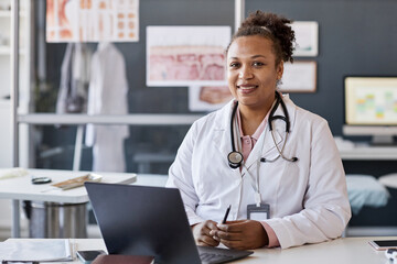 Portrait of smiling African American woman as female doctor working in clinic and looking at camera while sitting at desk with laptop copy space