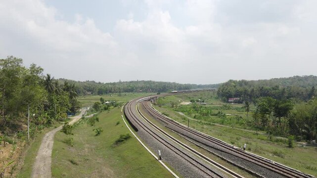 Drone shot, Indonesian train passing through a double track railway bridge winding on the left and right with rice fields, forests and villages.