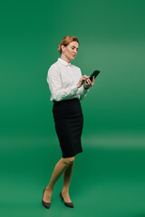 A professionally dressed woman stands poised, focusing on her smartphone against an eye-catching green background.