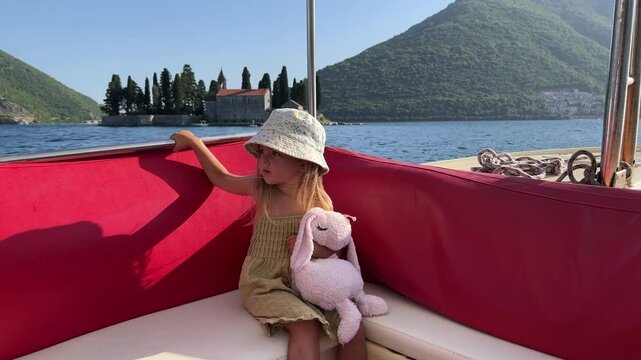 A young girl sits on a boat with a stuffed animal in hand, cruising on the water. The sky is clear, and she wears a hat for leisurely travel. Nearby trees add to the picturesque naval architecture