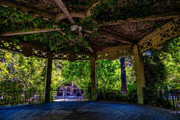 A wooden gazebo with intricate lattice work is adorned with lush green vines, creating a serene and...