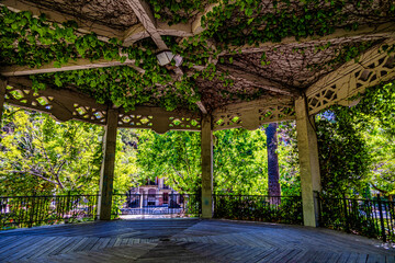 A wooden gazebo with intricate lattice work is adorned with lush green vines, creating a serene and...