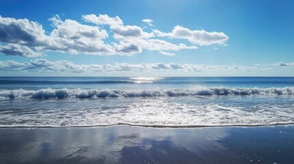 Fototapeta premium Tranquil ocean waves gently lapping at the shore under a bright blue sky with fluffy white clouds.