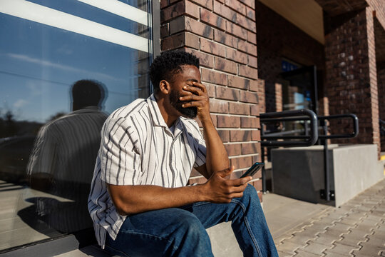 Disappointed black commuter sitting at bus station with cellphone in hands.