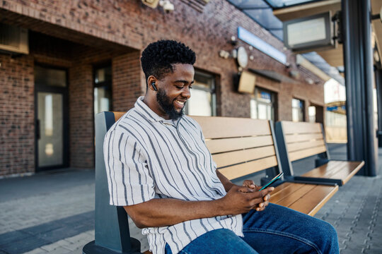 Happy black passenger sitting on bus platform with cellphone and headphones and waiting for bus.
