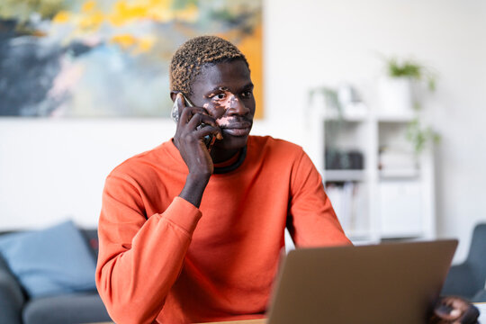 man with vitiligo wearing an orange shirt and sitting at a desk, smiling joyfully while talking on his phone, with colorful artwork in the background creating a warm, casual scene in a modern setting