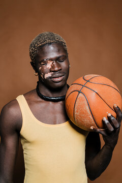 Black man with vitiligo is holding a basketball close to his face, wearing a yellow tank top and a black necklace, against a solid brown background with a calm and reflective expression.