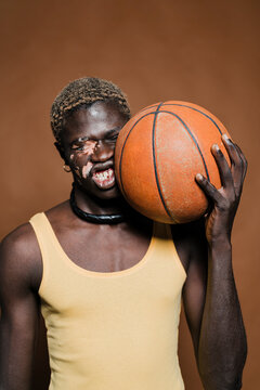 Black man with vitiligo poses with a basketball partially covering his face, wearing a yellow tank top and a black necklace, expressing quiet strength and uniqueness in a studio setting.