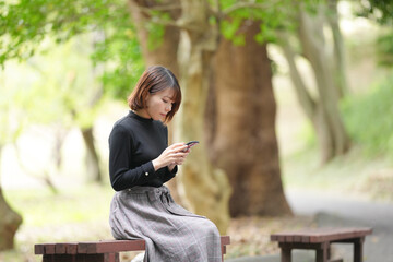 A young woman in her 20s with short hair wearing a black knitted long-sleeved shirt is sitting on a bench in the forest and operating a smartphone.
