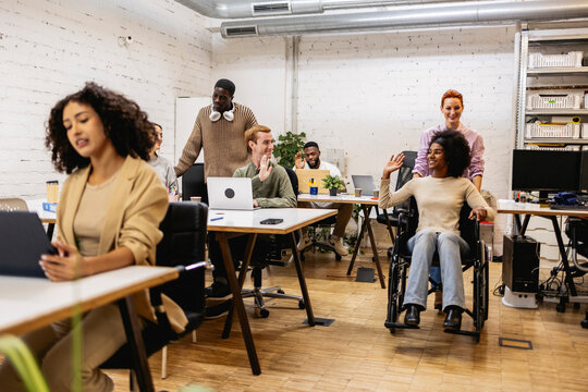 Diverse business team working in modern coworking space, woman using wheelchair greeted by coworkers