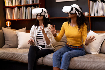 multiracial female friends enjoying virtual reality experience on cozy living room sofa, at home