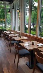 Obraz premium Modern restaurant interior with wooden tables and chairs, and large windows overlooking a green outdoor space.