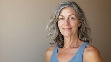 Fototapeta premium Mature woman in a blue tank top, calmly smiling, looking at the camera, warm beige background, soft daylight, medium shot