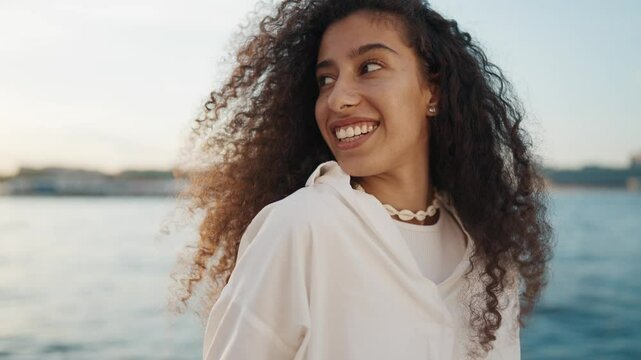 Attractive Arabic woman smiling broadly and viewing beautiful embankment. Medium portrait of joyful pretty lady with long curly hair, biracial female person, traveling to sea in summer vacation