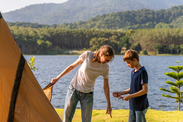 Fototapeta premium Mother and young son boy setting up a large, beige camping tent on a grassy area near a lake work together shared camping experience feelings of relaxation for a camping or outdoor adventure concept.