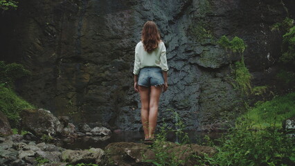 A young woman in a white shirt and denim shorts stands on a rocky outcrop near a waterfall in a lush, tropical forest in French Polynesia. The waterfall cascades down a dark, mossy rock face.