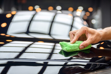 A hand applying ceramic coating with a green microfiber cloth to a glossy black car surface,...
