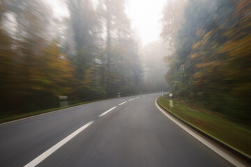 Fototapeta premium Long exposure, blurry autumn morning covered empty rural road through a forest in Europe. Wide angle view, limited visibility, bad driving weather concept, no people