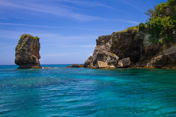 Fototapeta premium Gentle waves lap against rugged cliffs and a towering rock formation under a vibrant blue sky. Lush greenery tops the coastal landscape, adding a touch of vitality to the serene seascape.