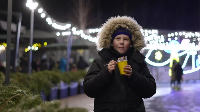Caucasian boy bites and chews fresh dessert pastry at winter Christmas market. Festive mood of child while snacking on treats in cold weather on Christmas Eve. Family leisure time in winter evening.