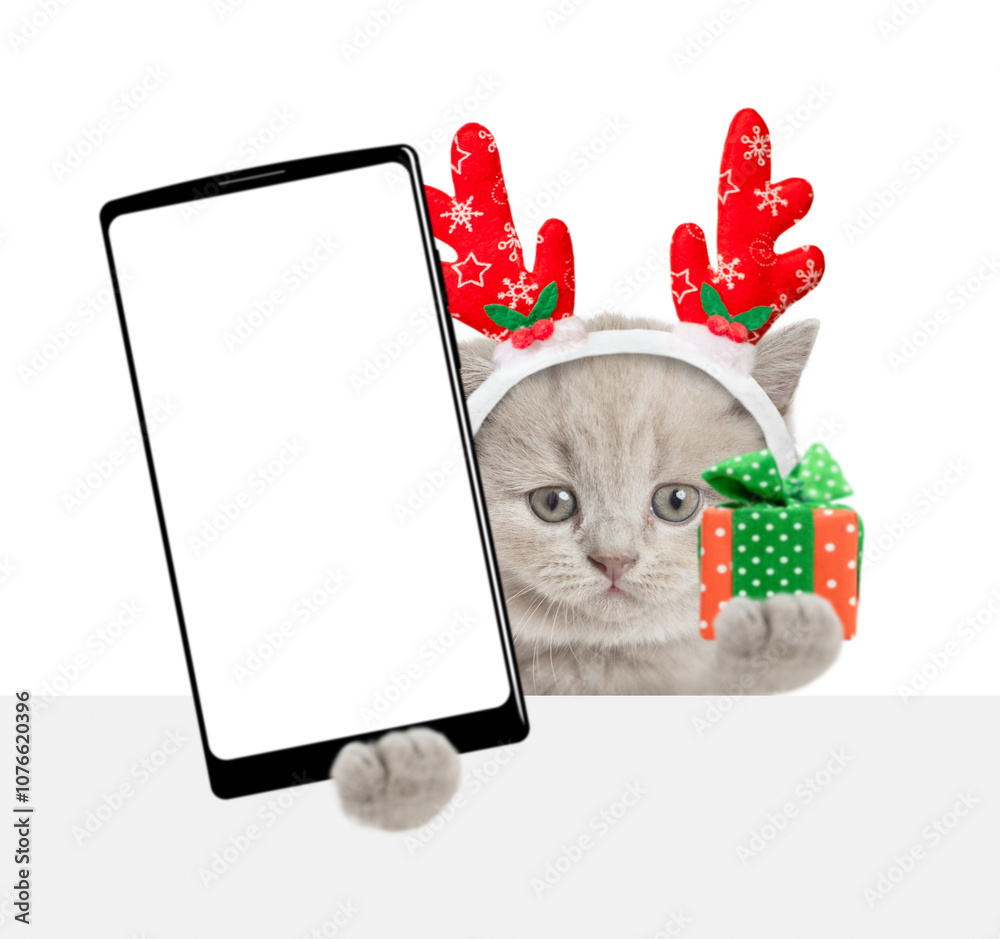 Poster cute kitten wearing red christmas hat holding gift box and showing smartphone with white blank scree