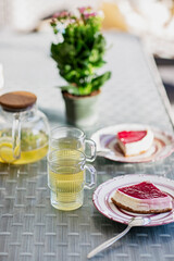 Piece of cheesecake with raspberry sauce and cup of green tea  Stil life of glass teapot with herbal tea lemon and meant and strawberry cheesecake on table outdoors vertical