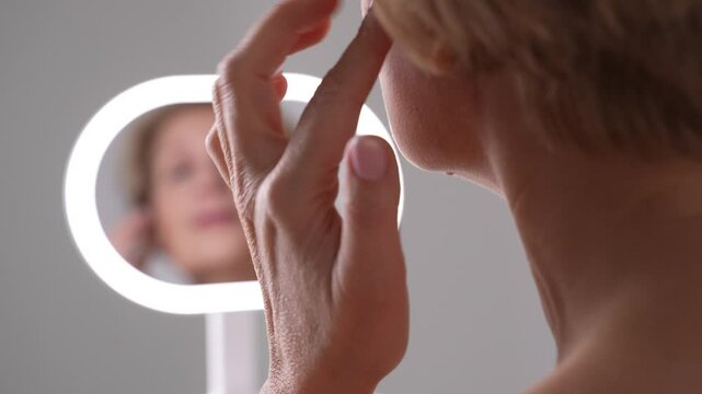 A confident middle-aged woman gently tapping her face with fingers as she looks in the mirror. Anti-aging nourishing skincare routine