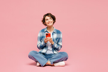 Full body young woman wear blue shirt white t-shirt casual clothes sits hold in hand use mobile cell phone look aside isolated on plain pastel light pink background studio portrait. Lifestyle concept.