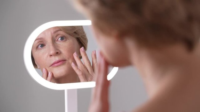 Morning skincare of a graceful middle-aged female, looking at skin changes due to aging with sadness. Anti-aging skincare, self-acceptance