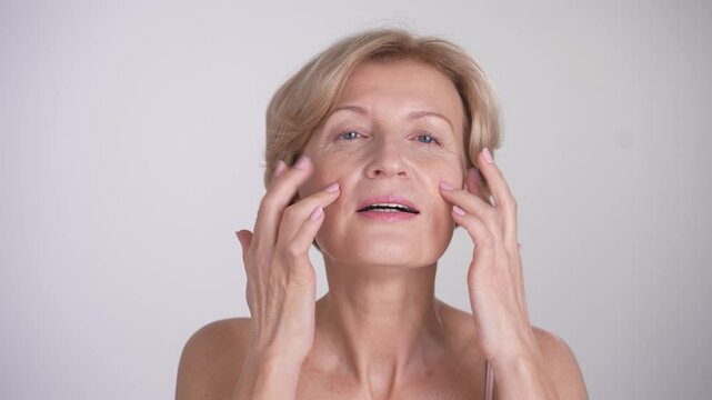 Portrait of a beautiful aging woman during her skincare routine. Positing happy female applying cream and finger-tapping. Graceful aging
