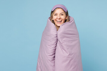 Calm smiling happy young woman wear purple pyjamas jam sleep eye mask wrapped in duvet rest relax at home look camera isolated on plain pastel light blue cyan background. Good mood night nap concept.