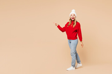 Full body side view merry young woman wear red warm sweater hat posing walking going point finger aside isolated on plain pastel beige background. Happy New Year celebration Christmas holiday concept.