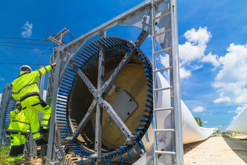 Two engineers in safety gear inspecting a wind turbine blade section on a construction site. They examine the metal framework with precision. Renewable energy project under a clear blue sky.