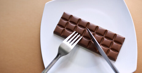 Fork, knife and chocolate with plate for unhealthy snack, sweet or cheat day above in studio on a...