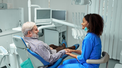 Dentist sits beside patient in dental chair, gently holding their hand and offering reassuring smile before procedure. Concept of health insurance, beauty, hygiene, dentist consultation. Ad.