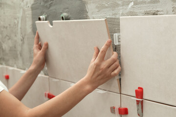 Woman tiling bathroom walls, applying adhesive on a tile and placing ceramic tile in right...