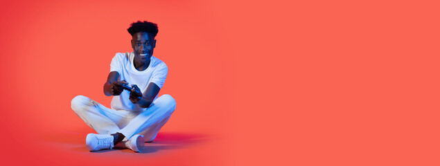 A cheerful young black man is seated with legs crossed, focused on playing video games. He is wearing a white shirt and jeans, enjoying his gaming experience against a vibrant orange background.
