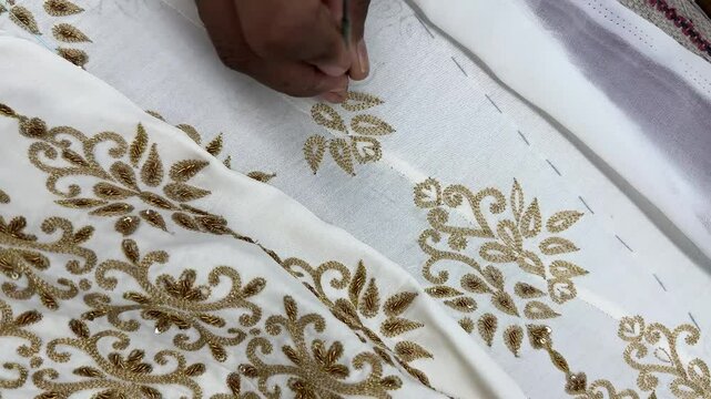 A Craftsman is doing hand embroidery on a white silk fabric, stretched in a wooden frame. Hand embroidery of a traditional motif with golden zari threads