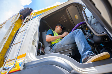 A truck driver wearing a reflective vest and cap looks at documents on a clipboard in the truck...