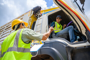 A logistics worker in safety gear hands over documents to a truck driver from outside the vehicle,...