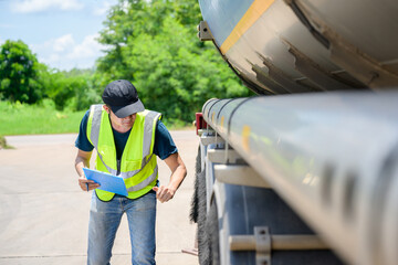 A safety inspector inspects the tires of a big fuel tanker while wearing a reflective vest. He maintains adherence to transportation safety regulations while holding a clipboard. © เลิศลักษณ์ ทิพชัย