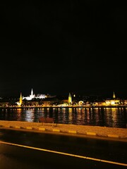 Obraz premium Nighttime view of a illuminated city skyline along a riverbank with reflections and historical architecture at dusk