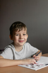 Naklejka premium A boy with blond hair in a white preschool T-shirt sits at a table in his room and writes numbers in a notebook with his left hand